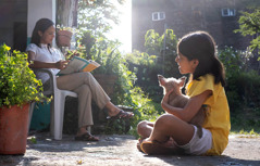 A young girl sitting on the ground and observing her mother, who is reading a book.