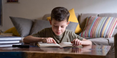 A young boy reading a book. There are a TV remote control and other books nearby.