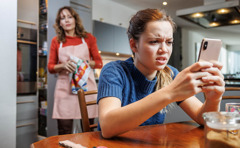 Una adolescente mira enojada su teléfono. Su mamá la observa a cierta distancia.