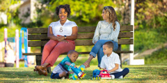 Deux mères de couleurs de peau différentes sont assises sur un banc dans un parc. Elles parlent en souriant pendant que leurs garçons jouent ensemble.