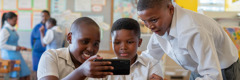 Okulda dikkatle telefonun ekranına bakan üç küçük erkek çocuk.
