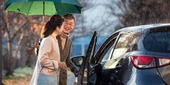年配の夫が妻のために傘を差しながら，車のドアを開けている。