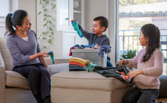 Una madre enseñándoles a sus hijos pequeños cómo clasificar y doblar la ropa. Su hijo le da la pareja del calcetín que ella tiene en la mano, y su hija dobla una camiseta.