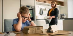 A frustrated boy struggling to assemble a small cardboard house. His mother patiently observes from a distance.