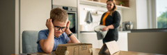 A frustrated boy struggling to assemble a small cardboard house. His mother patiently observes from a distance.