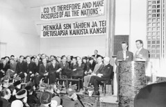 Irmãos e irmãs assistindo a um discurso do irmão Nathan Knorr que está sendo traduzido por um intérprete. Cartazes com o texto do ano de 1945 em inglês e finlandês estão pendurados na parede.