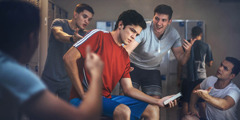 A teenage boy in a locker room, holding his phone while other teenage boys pressure him to make a decision.