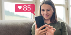 Një adoleshente buzëqesh ndërsa sheh celularin. Ka marrë 85 likes në llogarinë e saj në rrjetin social.