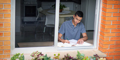 Um adolescente estudando em casa numa mesa perto de uma janela.