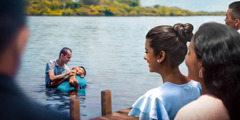 A man getting baptized in a lake as others watch from the dock.