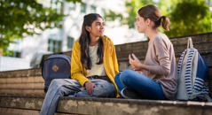 Two teenage girls looking at each other as they sit on an outdoor bench. One of the girls listens attentively as the other girl expresses herself.