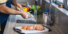 Mężczyzna myje nóż wodą i środkiem myjącym. Surowy kurczak i świeże warzywa leżą na oddzielnych deskach do krojenia.