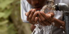 A young boy using his hands to drink water from an outdoor spigot.