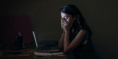 Eine junge Frau sitzt mitten in der Nacht aufgewühlt vor ihrem Computer und vergräbt ihr Gesicht in den Händen.
