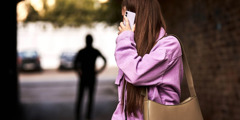 A criminal stalking a woman who is alone. She sees the criminal behind her and uses her mobile phone to call for help.