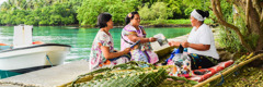 Two of Jehovah’s Witnesses offering a magazine to a woman who is sewing by the shore.