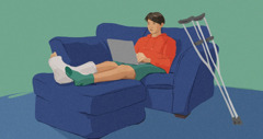 A teenage boy with a cast on his leg, sitting on a sofa while using his laptop.