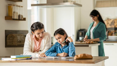 Uma adolescente ajuda a irmã mais nova a fazer os trabalhos de casa enquanto a mãe prepara uma refeição na cozinha.