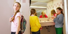 A teenage girl glances at other girls standing in front of a mirror putting on makeup