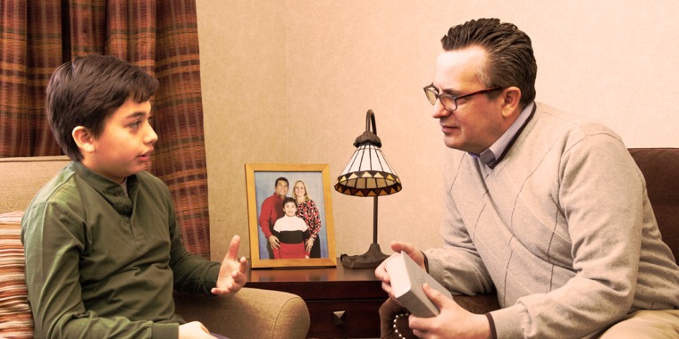 A boy sits beside a picture of his family and talks with a man who is holding a Bible
