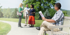 Мъж държи трактат и наблюдава как една жена говори със Свидетели на Йехова до подвижния им щанд