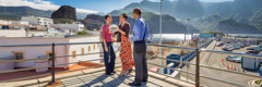Two of Jehovah’s Witnesses speaking to a young woman on an elevated walkway that affords a view of the coastal town and mountains.