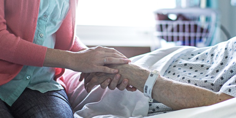 A young woman holds an older person’s hand in a hospital