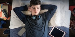 Ein Jugendlicher, der von Unterhaltungstechnik umgeben ist, liegt auf dem Bett und starrt an die Decke