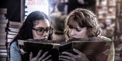 Two teenage girls look at an old book that features the occult