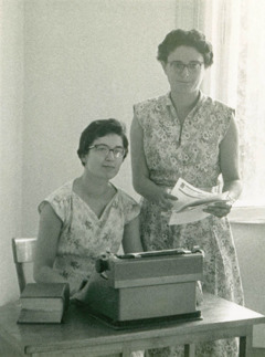 Irma sentada em frente a uma máquina de escrever com Ilaria em pé ao lado dela.