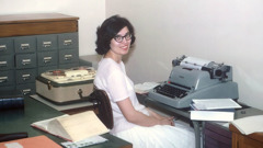Irma está sentada frente a una máquina de escribir. A su lado tiene una grabadora de carrete.