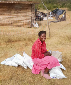 Prisca outside her home, smiling with several bags of food supplies.
