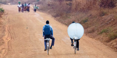 Brothers riding bicycles on a dirt road, transporting satellite equipment to their congregation.