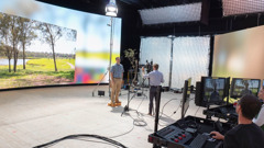 A brother in a studio, standing in front of a video wall displaying a parklike setting. Other brothers calibrate the equipment.